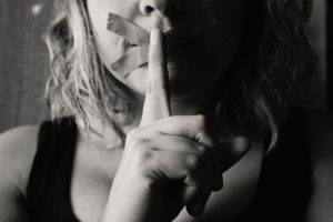 Woman with tape covering her mouth to silence her and a finger to her lips indicating she's keeping secrets.