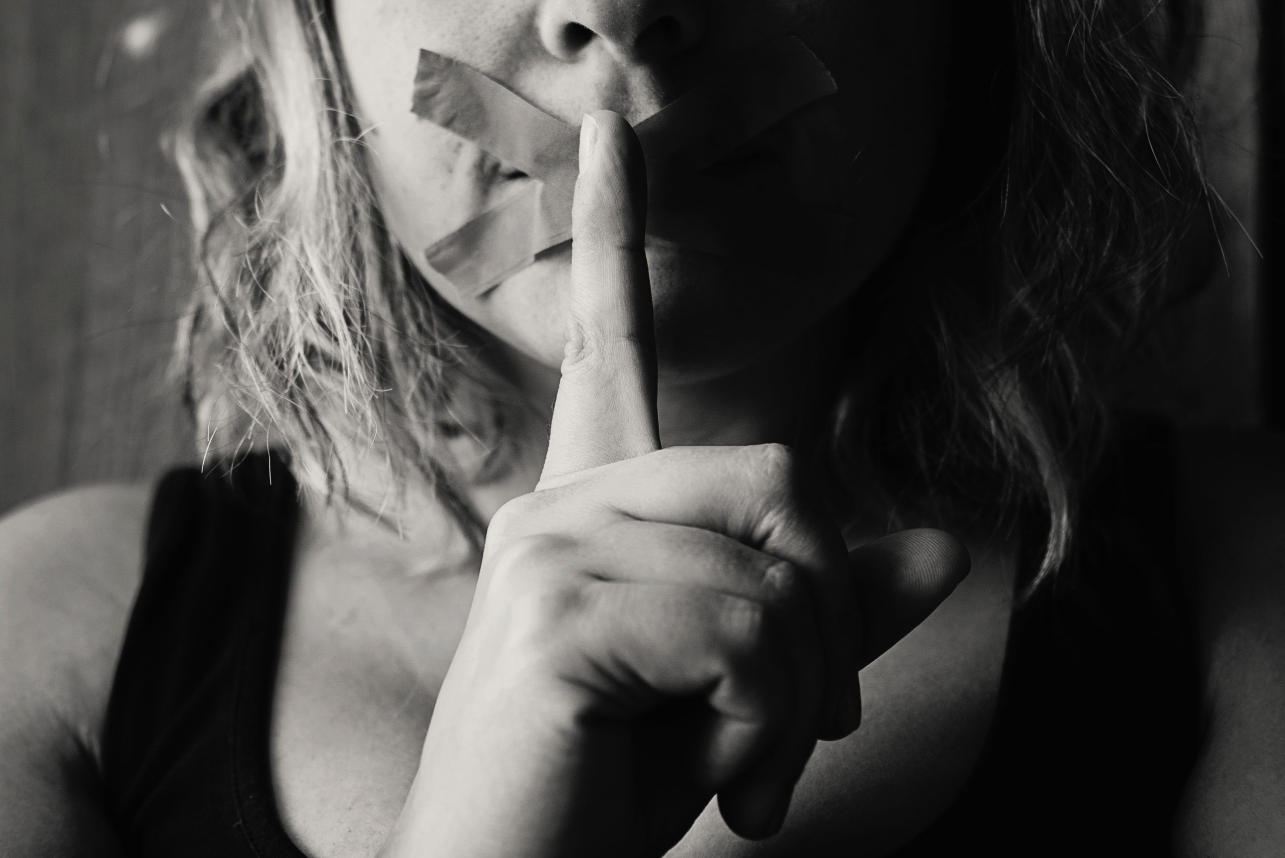 Woman with tape covering her mouth to silence her and a finger to her lips indicating she's keeping secrets.